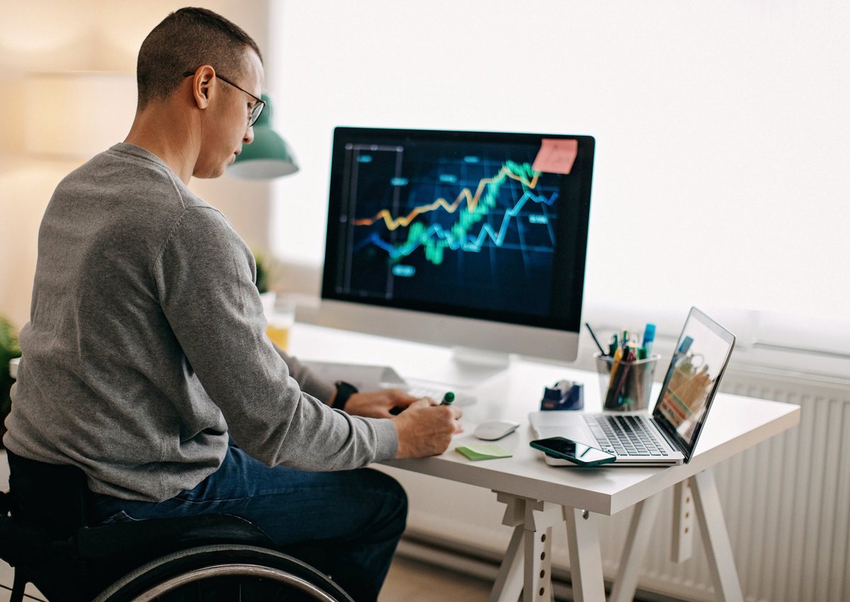 A person using a wheelchair analyzes a stock market graph on their desktop computer and takes notes.