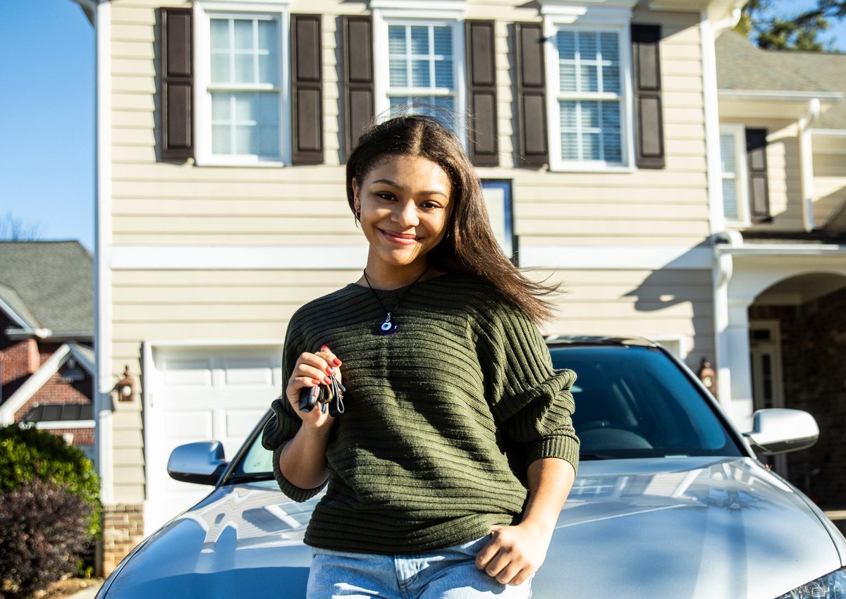 Young girl poses with keys in front of new car parked in front of a house.