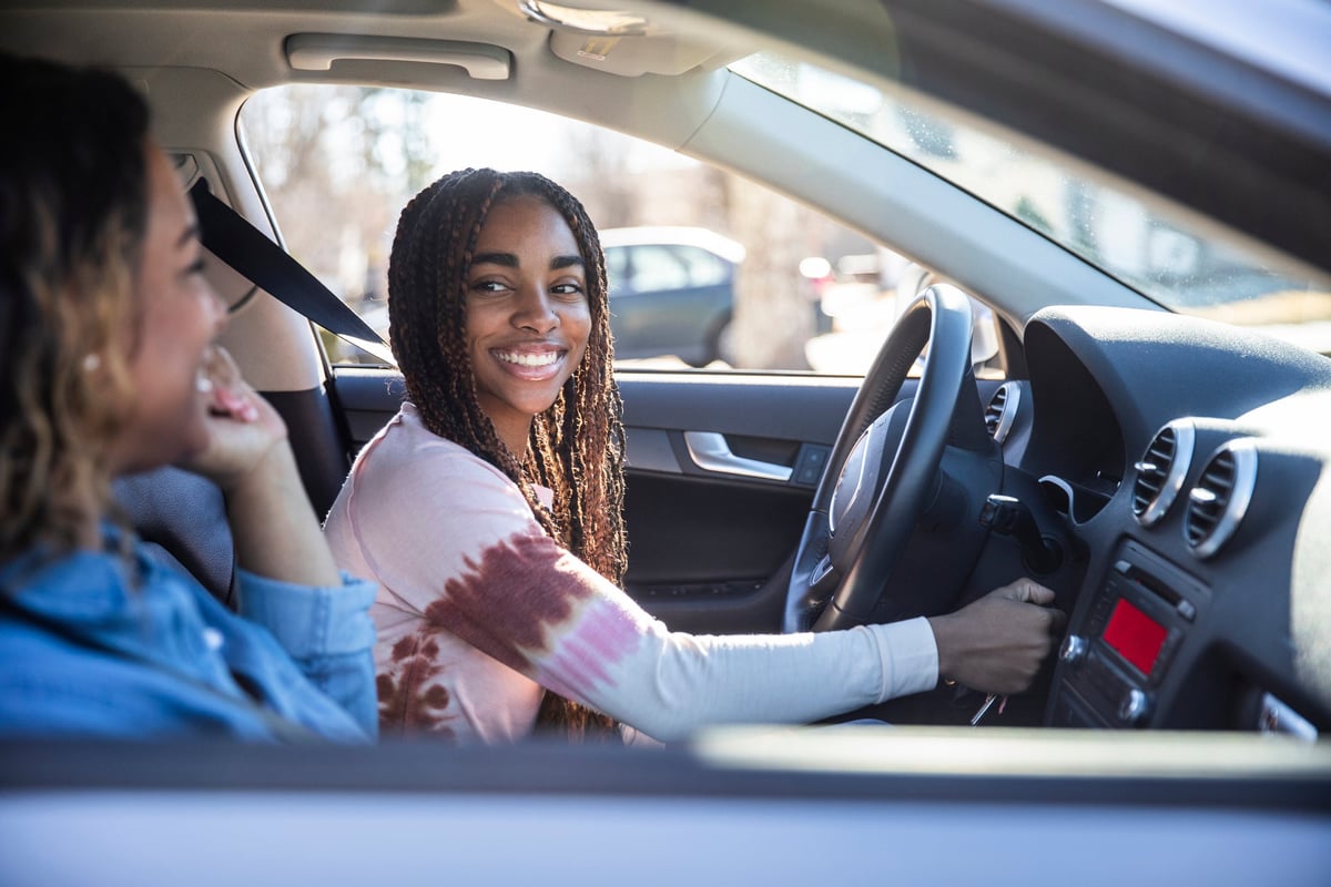 A teenager smiles at their mother as they turn on the car ignition.