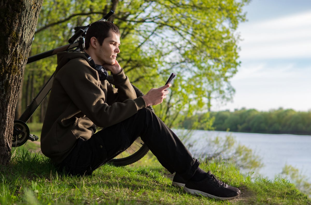 A person sitting on the riverbank next to his bicycle and looking at his smartphone.