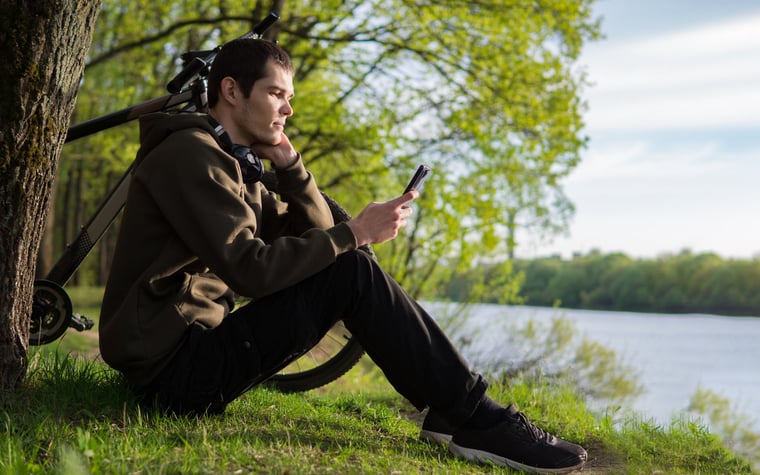 A person sits on the bank of a river next to his bicycle, looking at his smartphone.