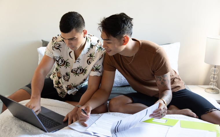 A couple sits on their bed looking at a laptop and paperwork.