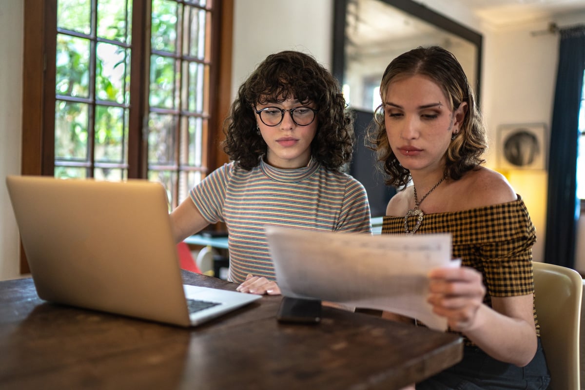 A couple discusses finances at the kitchen table using a laptop and paperwork.