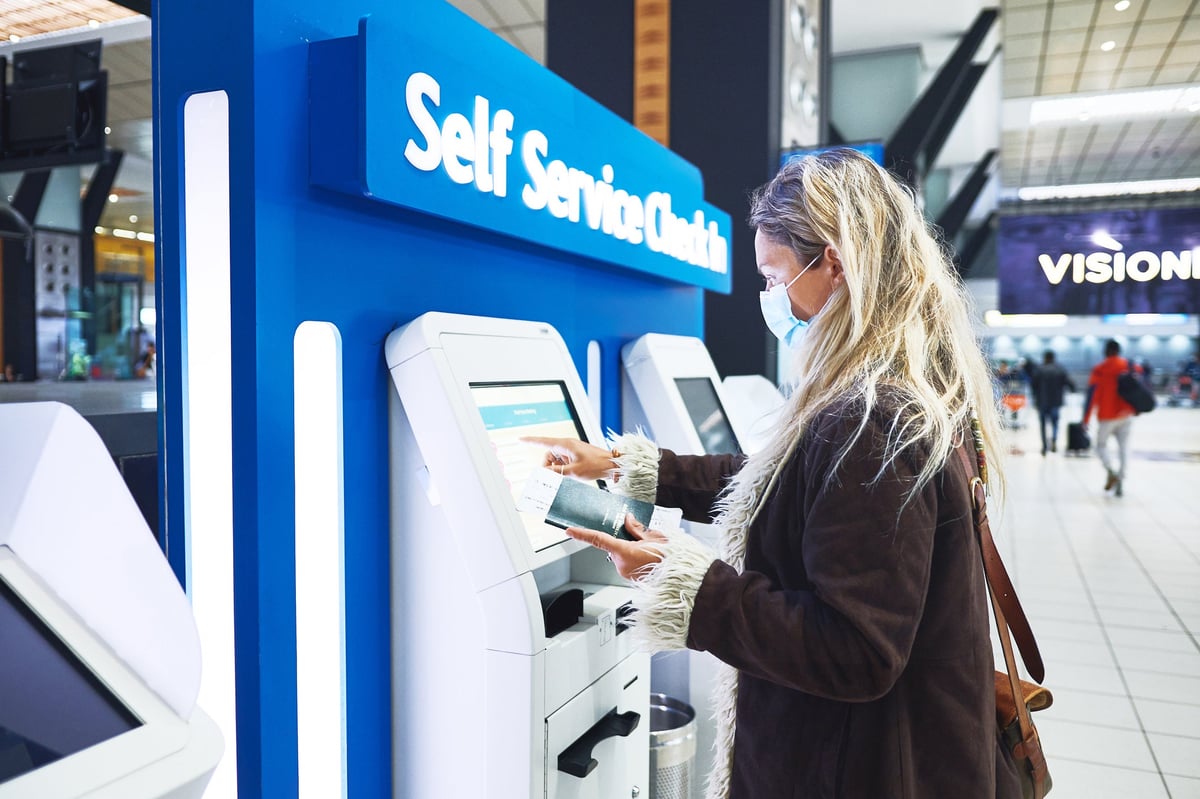 A young adult wearing a facemask uses a kiosk to check in for a flight.