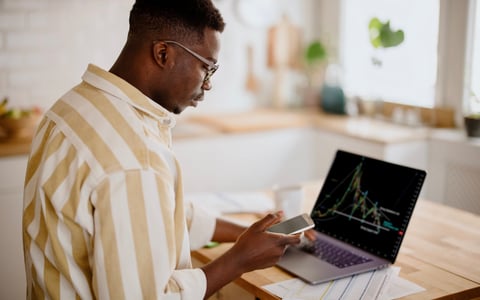 A person analyzes a cryptocurrency graph on their laptop while looking at other information on their smartphone in their kitchen.