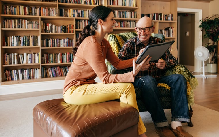 A caregiver holds up a tablet and laughs with their older parent.