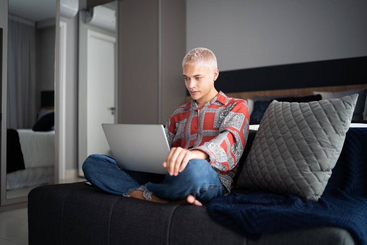 A young adult sits cross-legged and types on a laptop in his bedroom.