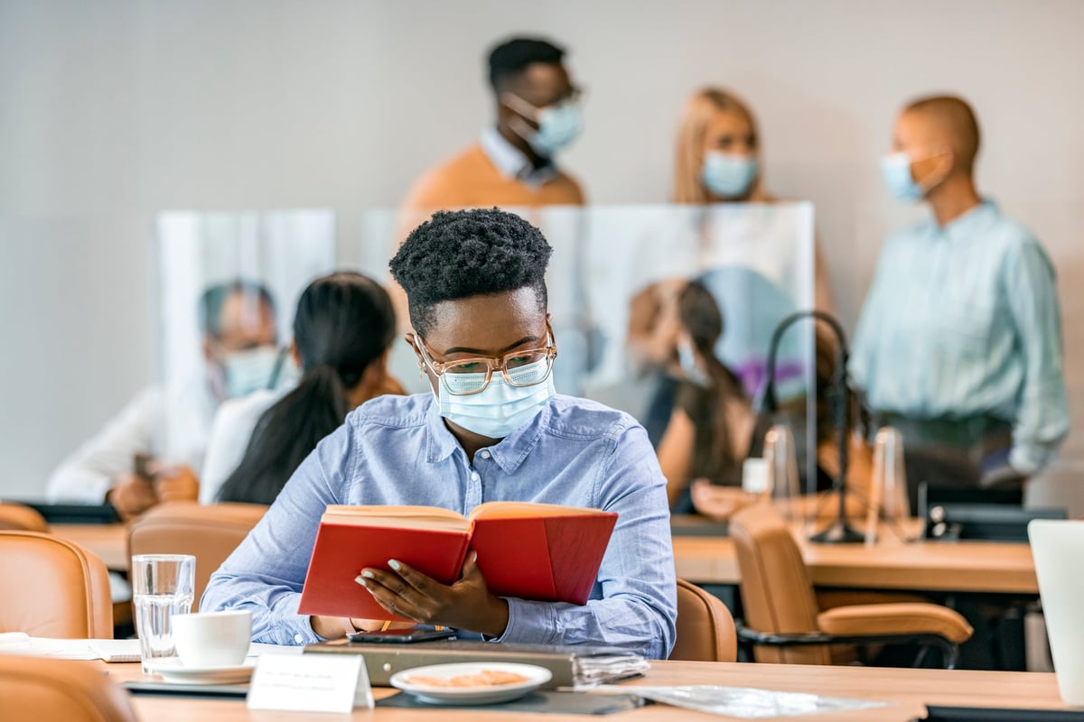 A businessperson reads a research book while wearing a mask in an office cafeteria.