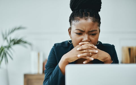 A young business professional worriedly looks at a computer screen as their head rests on their clasped hands.