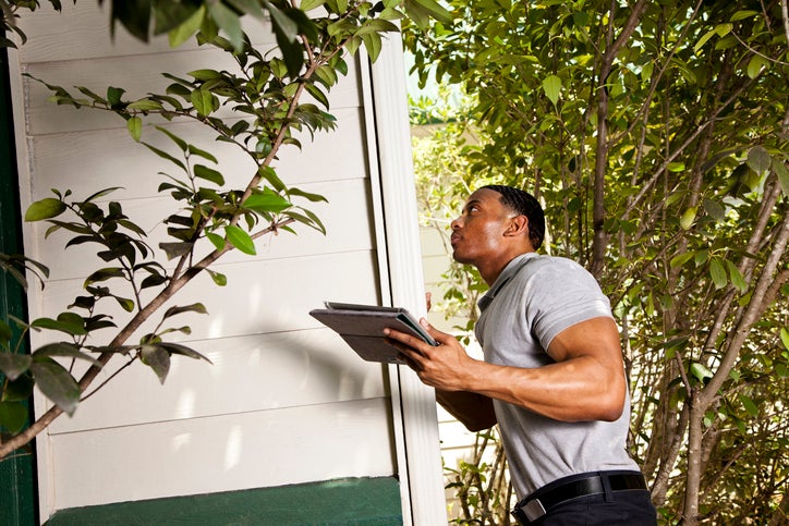 Home inspector checks the exterior walls of a house and uses a tablet to record results.