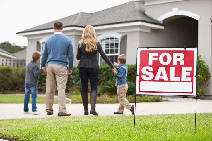 A family looks excitedly at a home with a For Sale sign on the lawn.