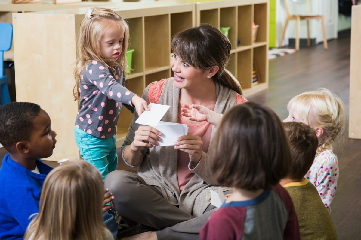 A pre-school leader sits with a group of children in a classroom, holding up flash cards.