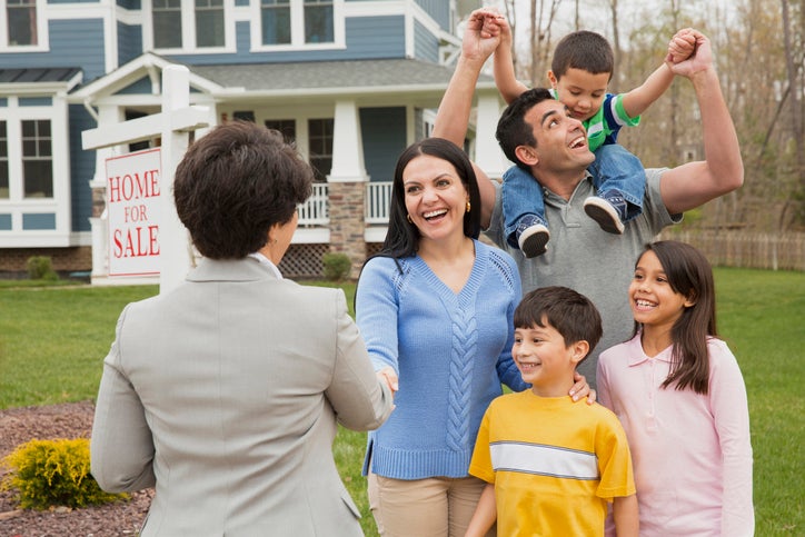 A parent shakes hands with a realtor as their family celebrates. A house with a sign that reads "Home for Sale" is behind them.