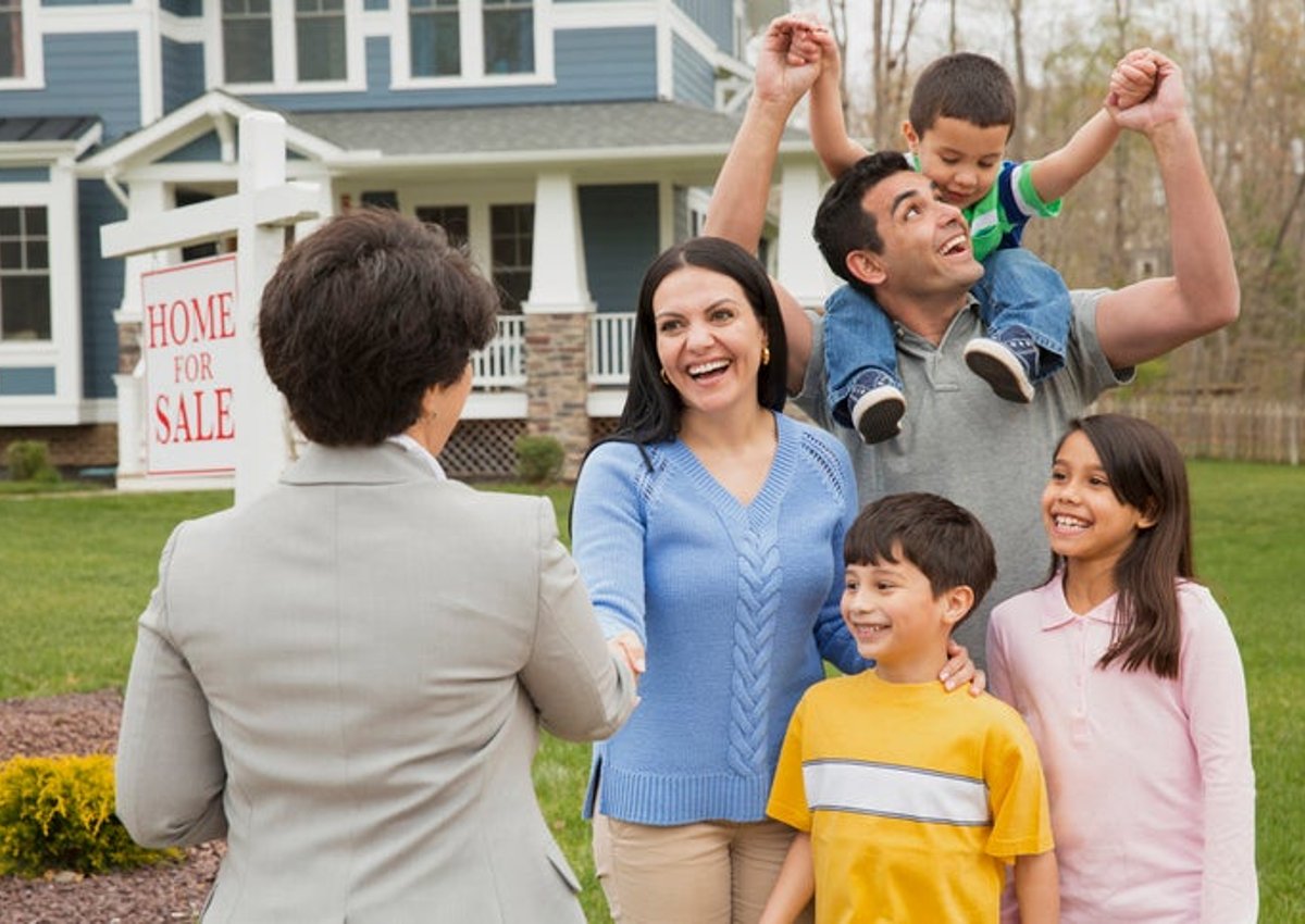 A parent shakes hands with a realtor as their family celebrates. A house with a sign that reads "Home for Sale" is behind them.