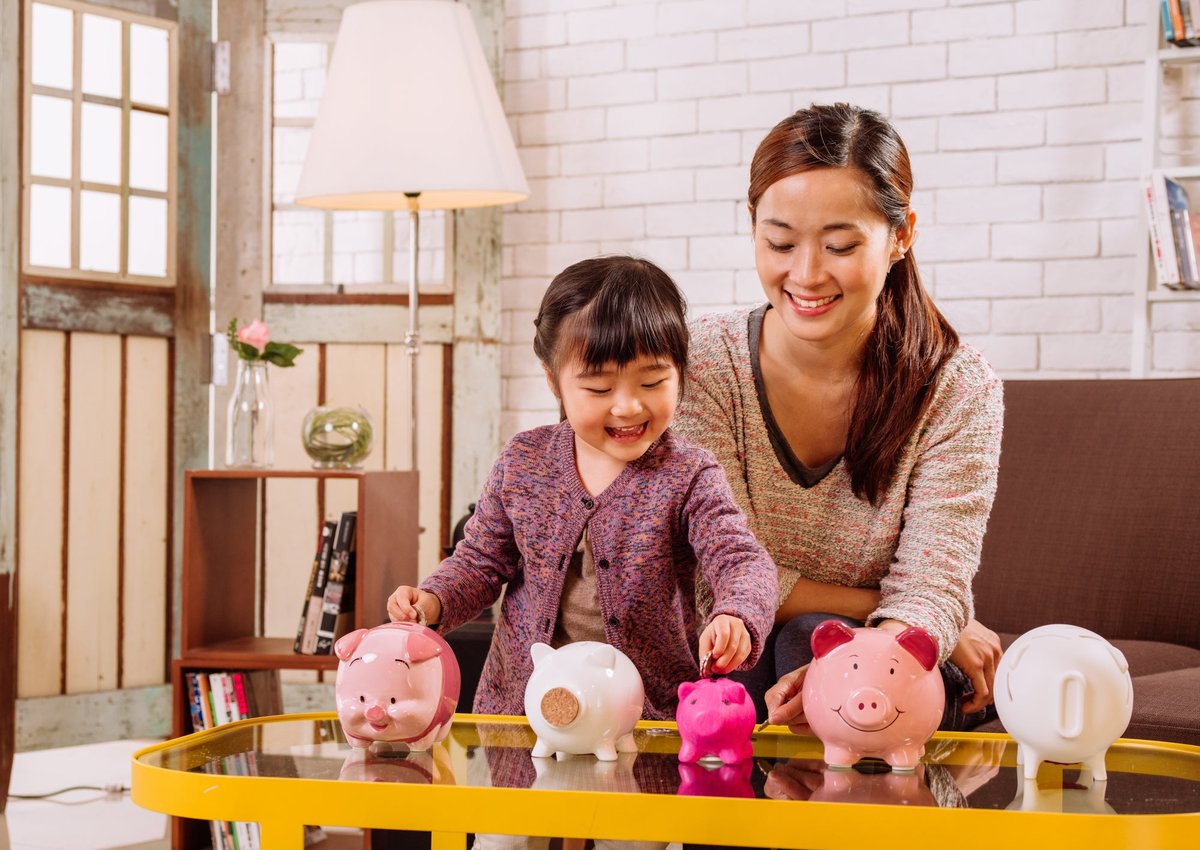A child puts coins into differently sized piggy banks under the happy supervision of her parent.
