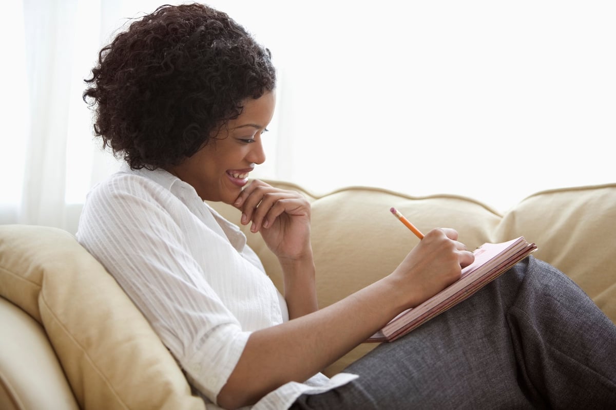 A person reclines on their couch while writing with a notepad.