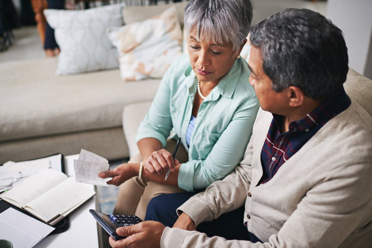 Un couple plus âgé utilise un ordinateur portable, une calculatrice et des papiers pour établir son budget tout en étant assis sur le canapé du salon