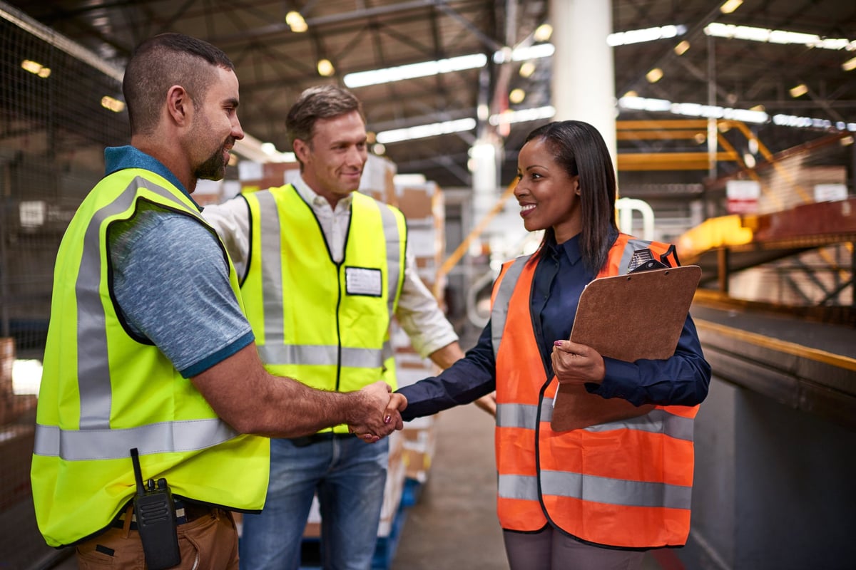 Two employees wearing work vests shake hands in a warehouse while another worker smiles.