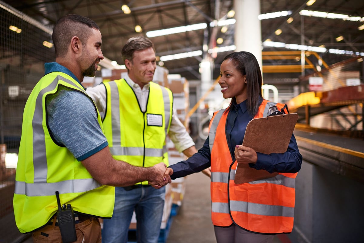 Two employees in work vests are shaking hands in a warehouse while another is smiling.