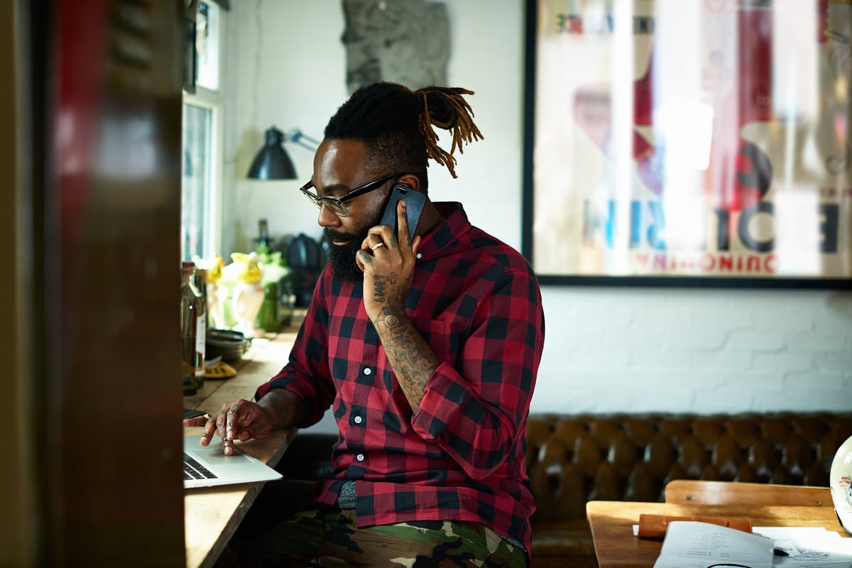A person with a beard and tattoos talks on a smart phone while using a laptop in a design studio.
