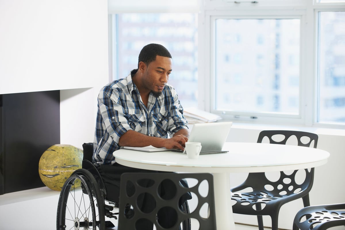A person using a wheelchair types on a tablet while sitting at a table in a public space.