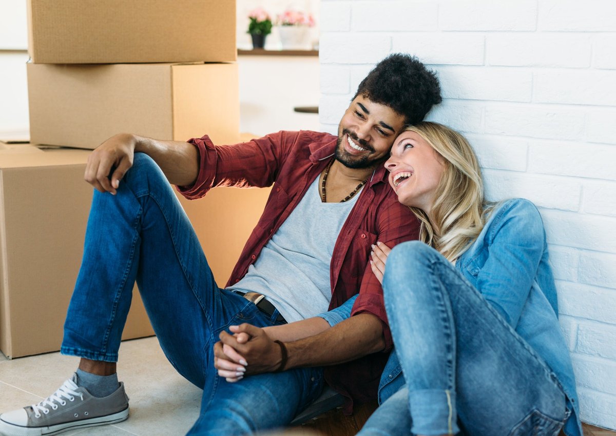 A young couple holds hands and smiles as they sit on the floor near moving boxes.