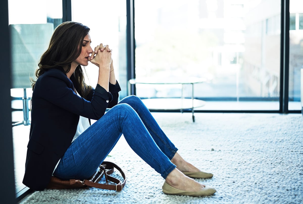 A stressed person holds their clasped hands to their lips while sitting on the floor of an office.