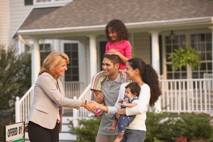A family with children shakes hands with a real estate agent in front of a house.