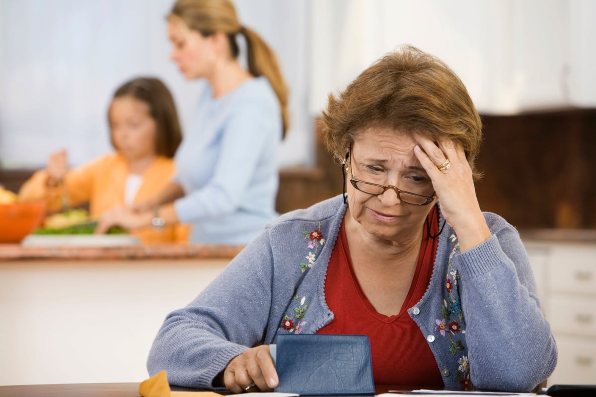 An older person worriedly reviews their checkbook in the kitchen while their family cooks in the background.