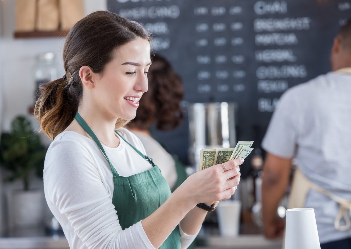 A smiling young barista counts cash in coffee shop.
