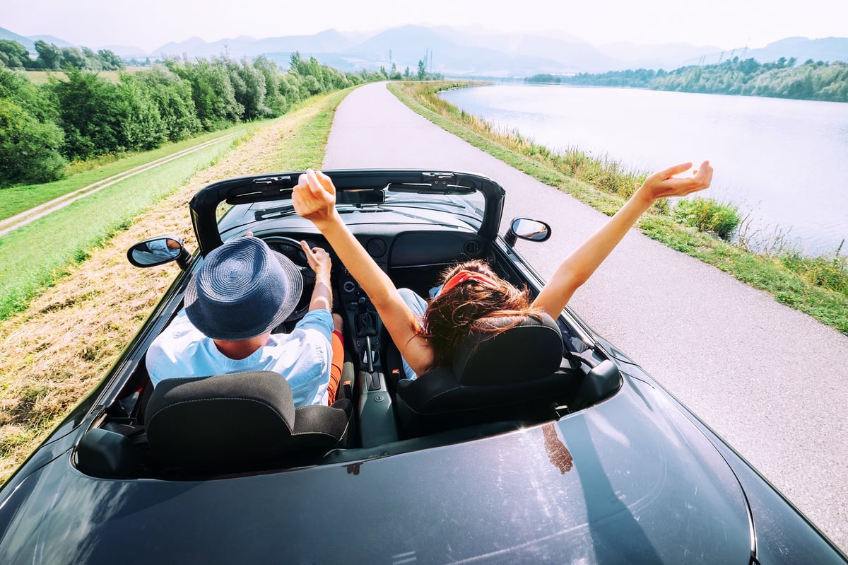 young man and woman riding in a convertible next to a lake on a sunny day.