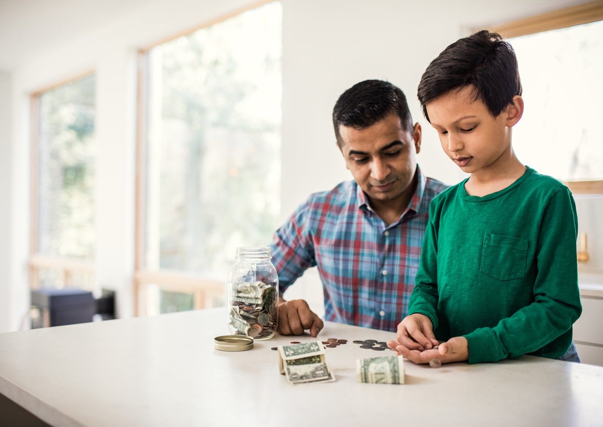 A parent and child count dollar bills and coins from a jar in their kitchen.