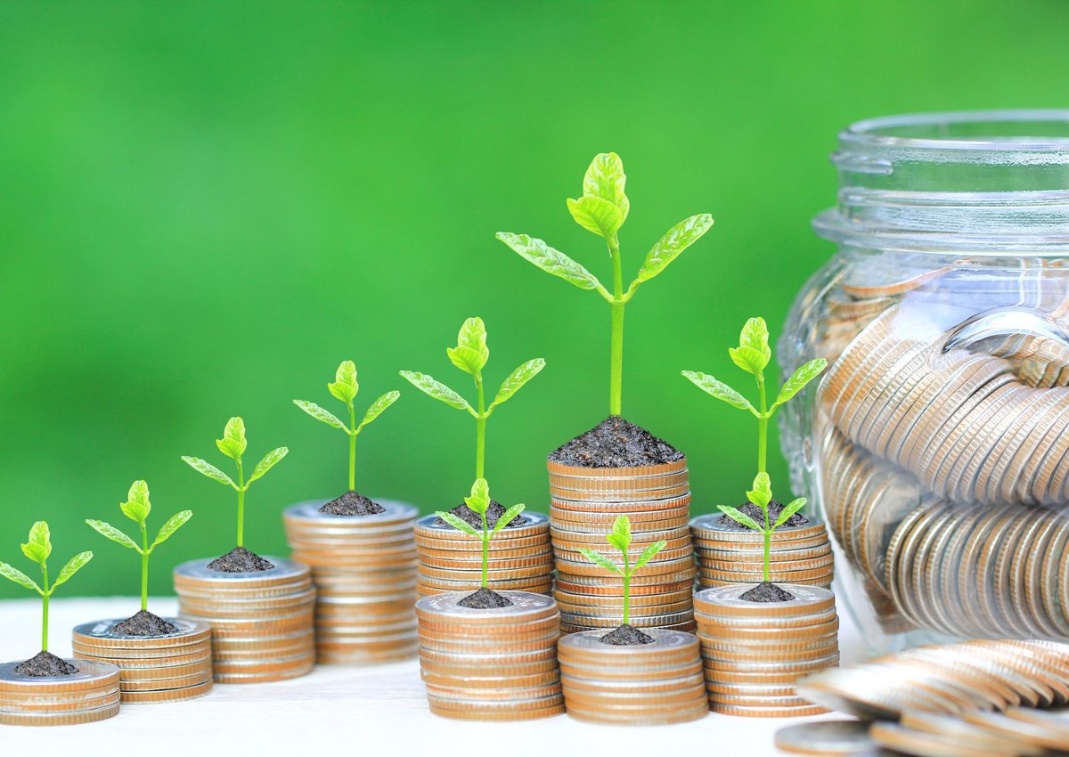 Tiny sprouts growing out of coin stacks of varying heights beside a jar full of coins.