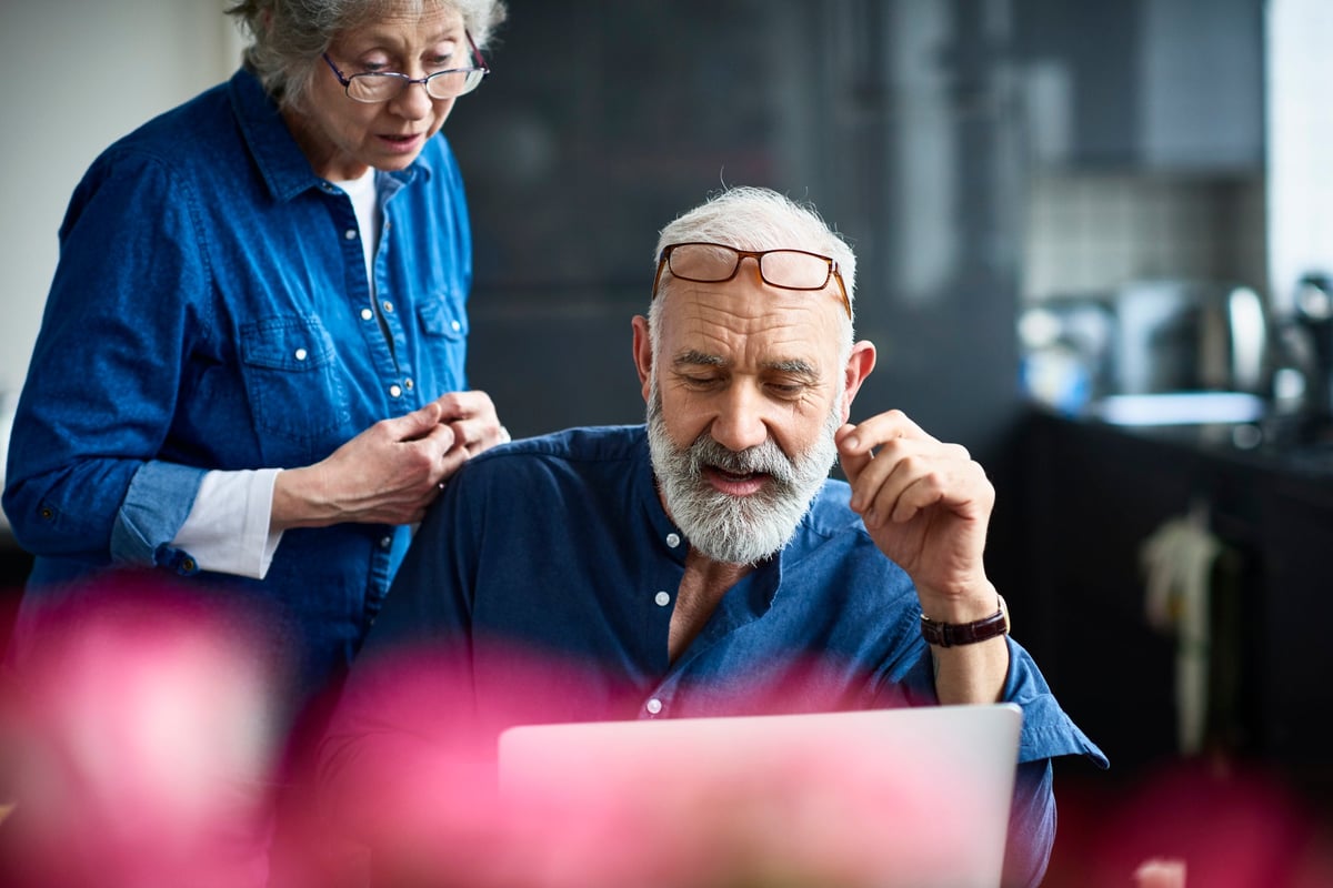 An elderly couple are discussing something on a laptop screen.