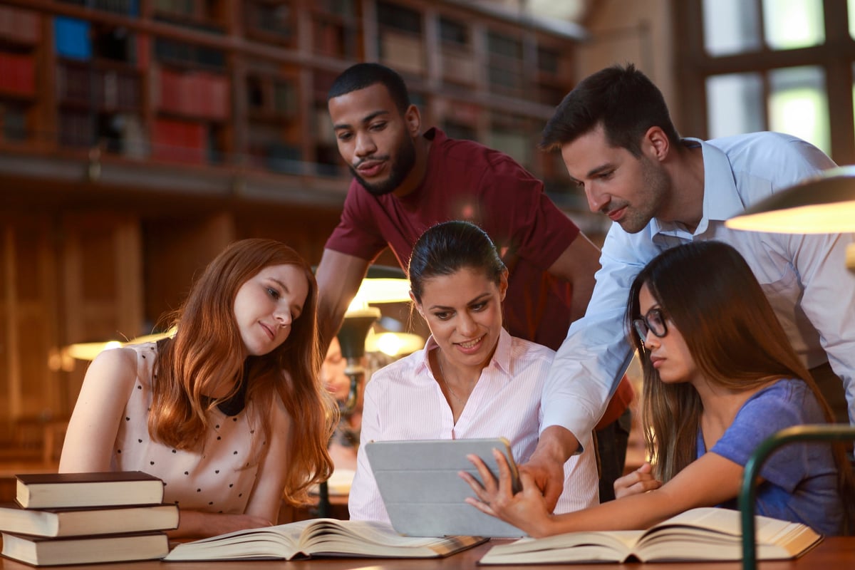 A group of young people gather in the library and look at a tablet.
