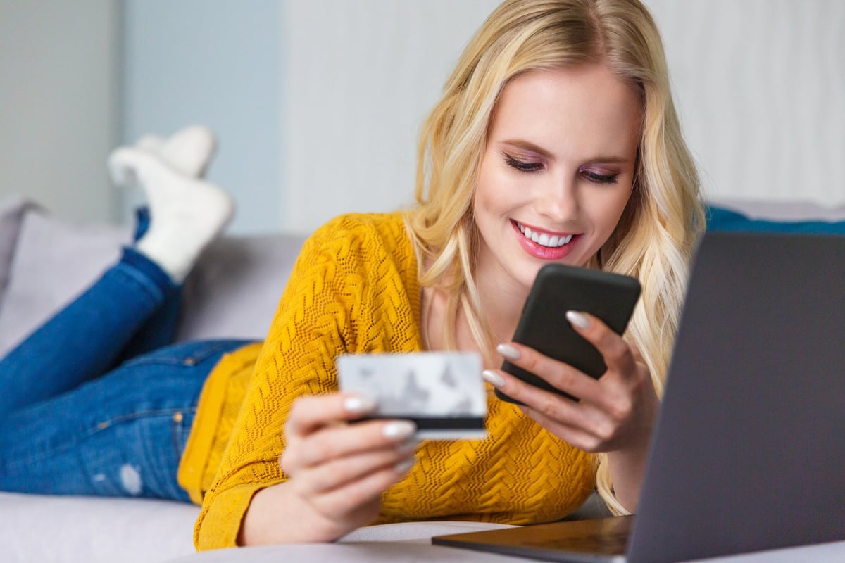Smiling woman lying on her couch, using her cell phone, and holding her credit card.