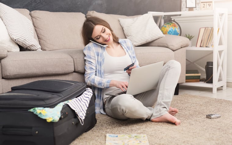 Woman sitting on floor beside suitcase talking on her cell phone and using a laptop.