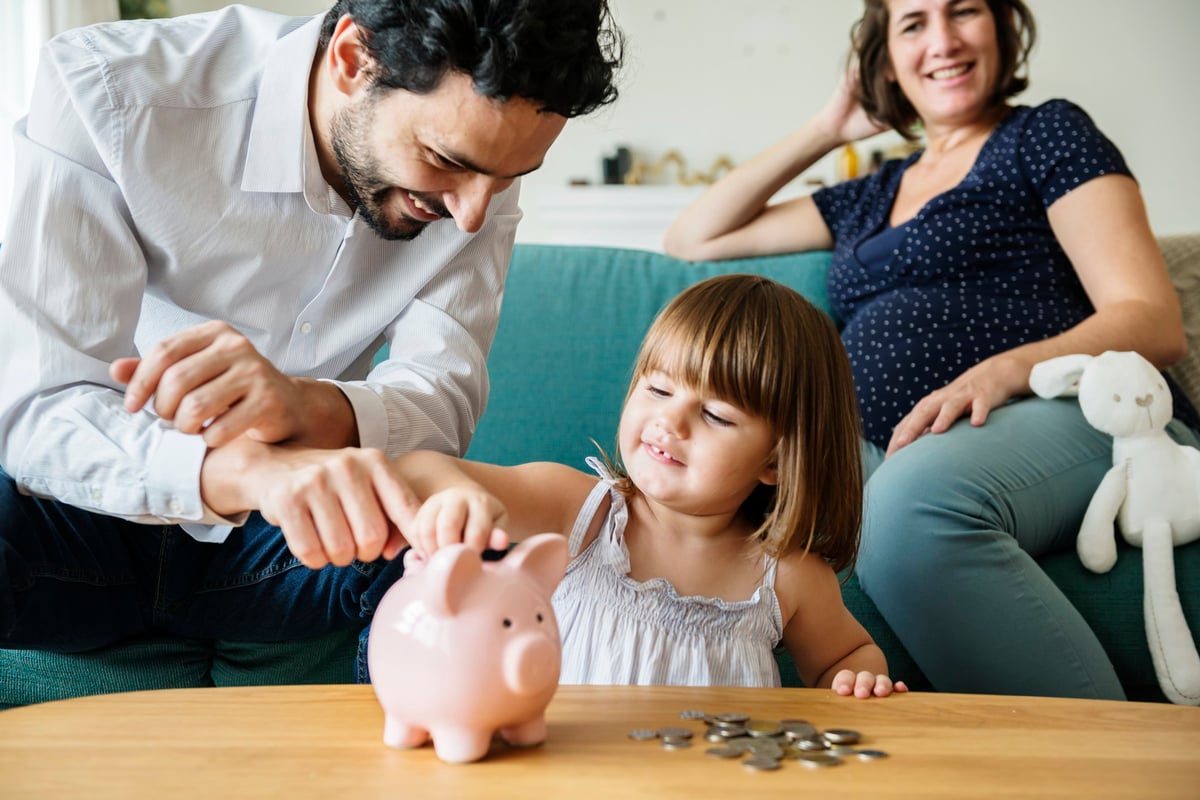 Happy Father Showing His Daughter How To Put Coins In Piggy Bank While Mom Watches