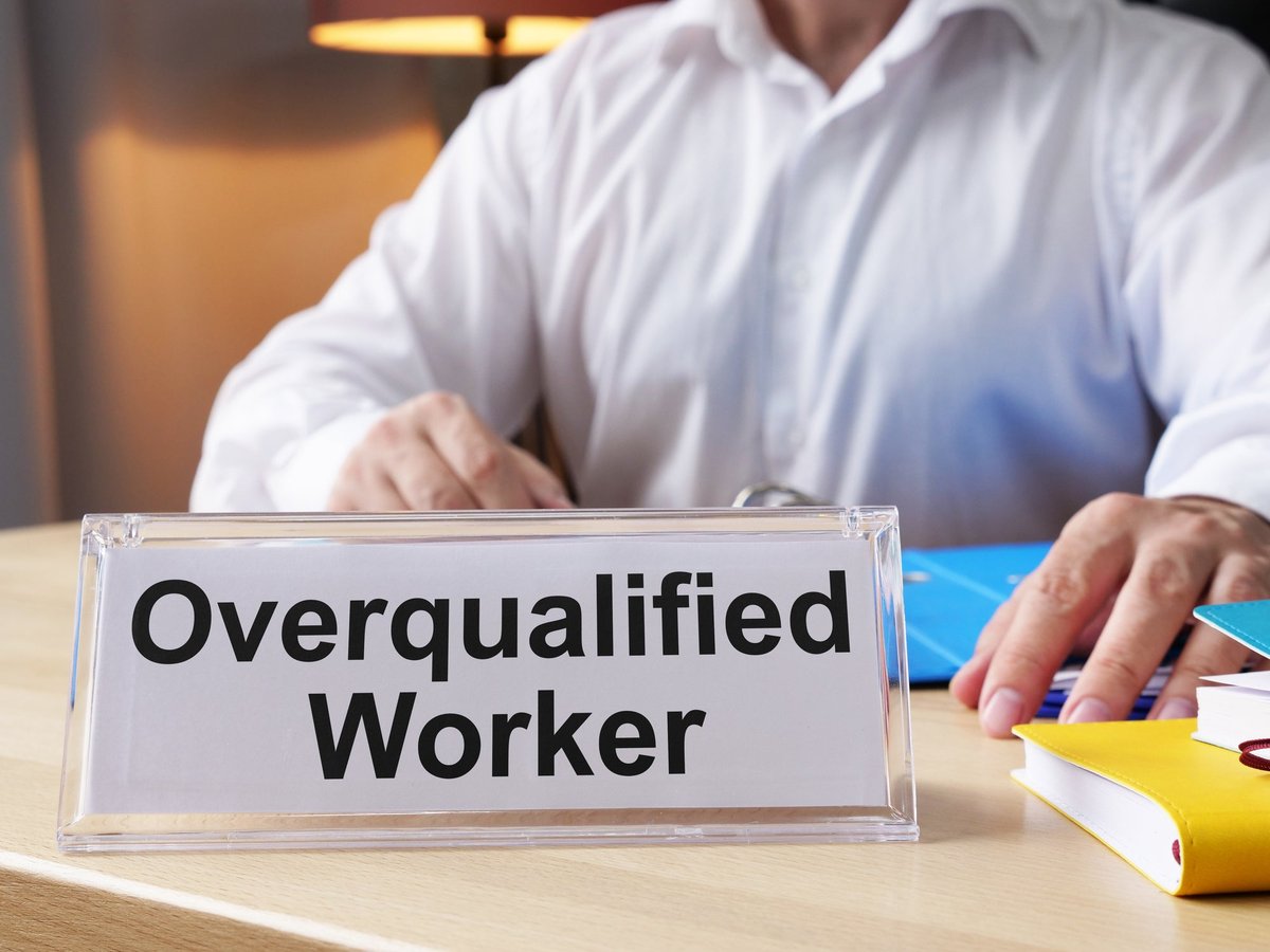 A man in the background sits at a desk with a name plate that reads "Overqualified."