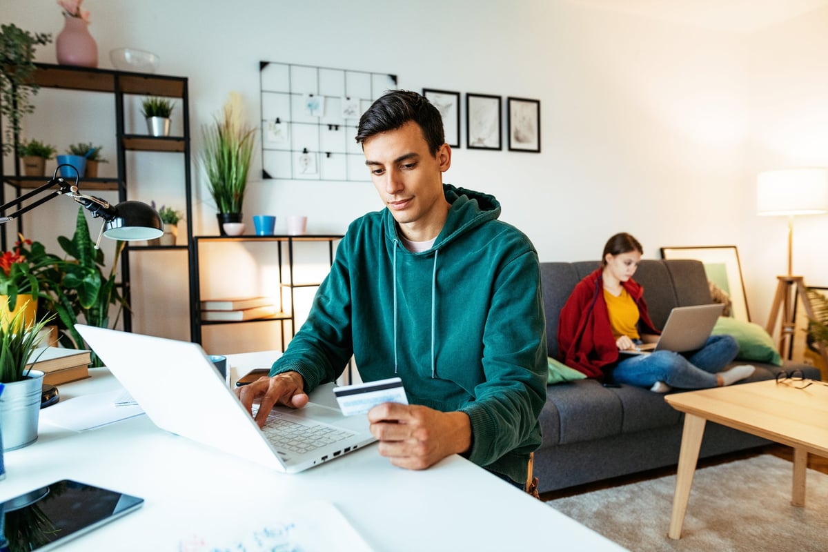 At home, a man sits at a table and is using his computer while holding his credit card.