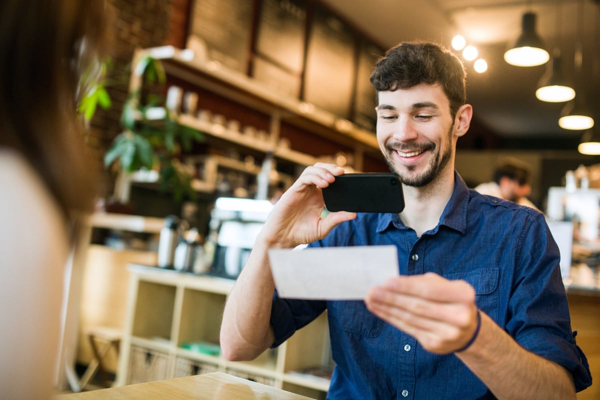 A man sits in a cafe and uses his phone to deposit his check.