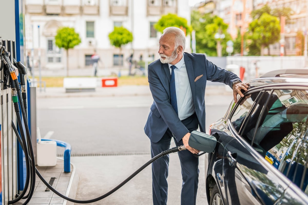 An older man in a suit fills up his car with gas.