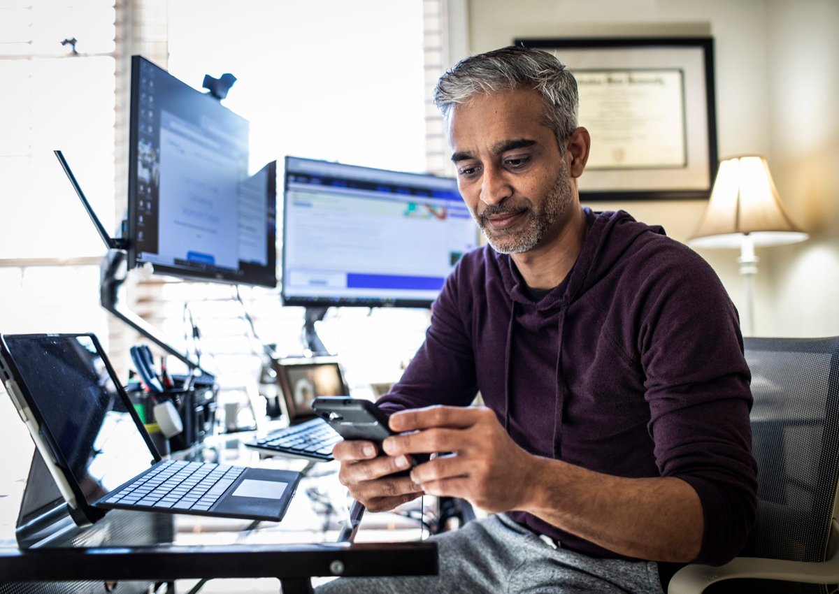 A man sits in a home office with two computers and a laptop. He is looking at his phone.