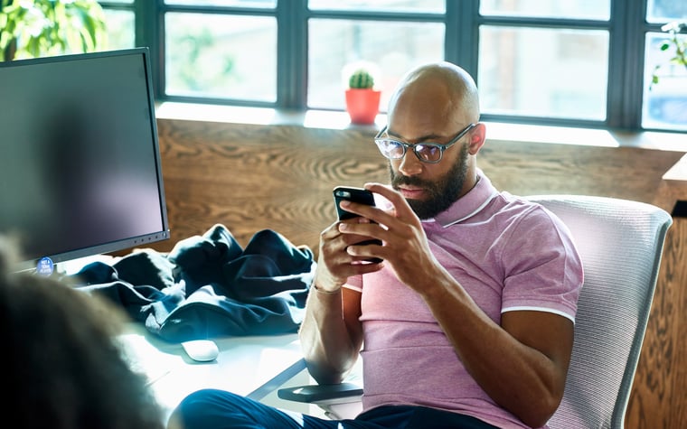 A person sitting at a desk looking at a cell phone with a pensive expression.
