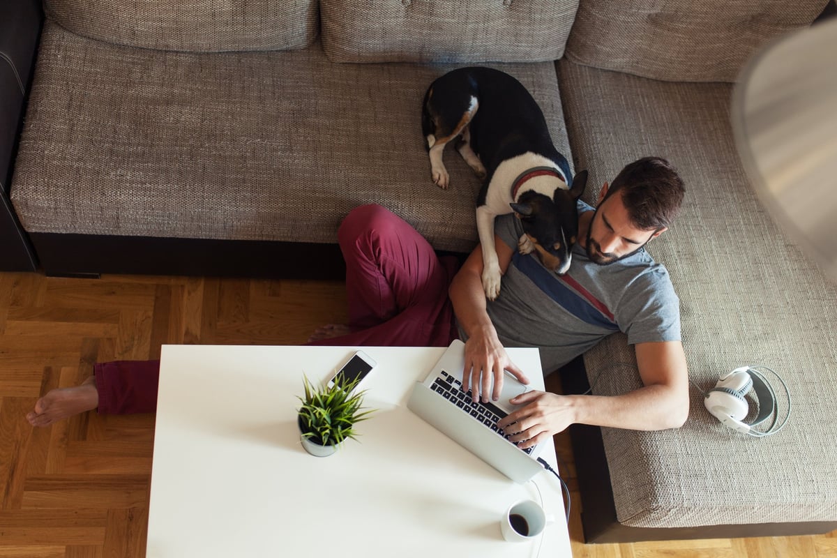 A man on a laptop sitting on the floor next to his couch with his dog's head on his shoulder.