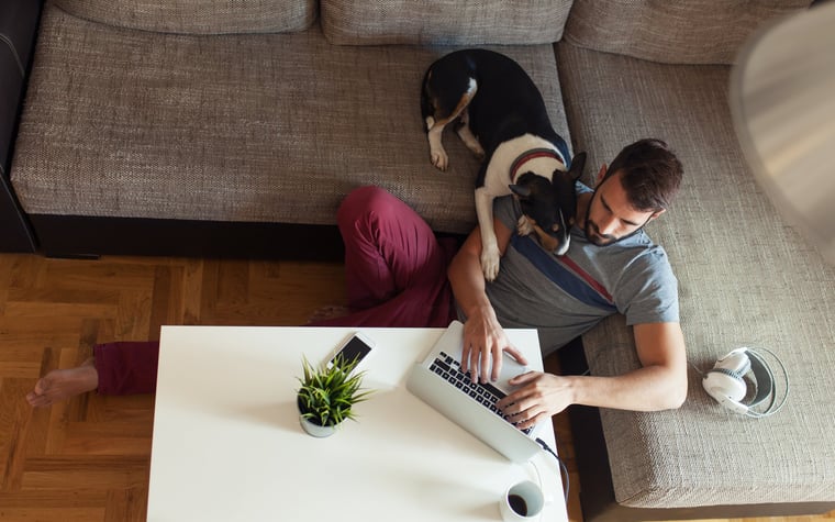 A man sits on the floor in front of his sofa with his dog looking over his shoulder at his open laptop.