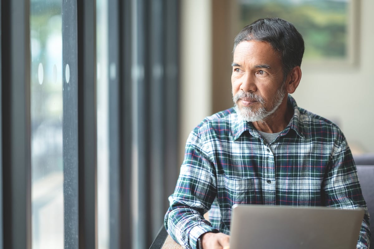 A middle-aged man holding a laptop and looking out of a cafe window.