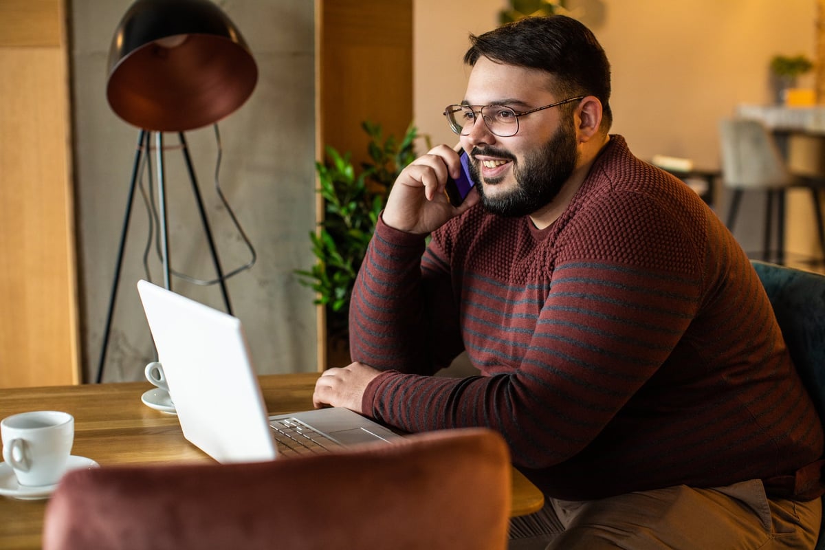 A man talks on the phone while sitting in front of his laptop in a coffeeshop.