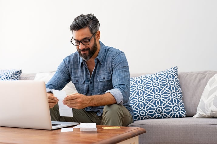 A man looking through paper bills while sitting on a couch in front of a laptop.