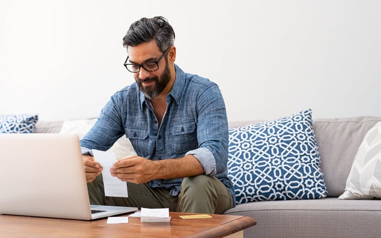 A man looking through paper bills while sitting on a couch in front of a laptop.